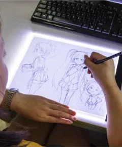 Una persona dibuja en una tableta de luz, con la mano derecha sosteniendo un lápiz y creando personajes estilo anime, mientras un teclado se encuentra en el fondo, y en la muñeca lleva un reloj, resaltando el uso creativo del Almohadilla Led Diamond Painting A3/A4/A5.