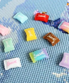 Pequeños objetos cuadrados y brillantes con la palabra "Sweet", en colores pastel sobre un fondo de mosaico azul claro, forman parte del diseño Cover Minder Diamond Painting​ Candy Crush.