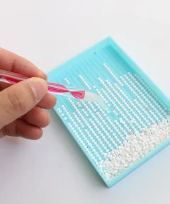 Una mano sostiene un aplicador con mango rojo y transparente sobre una bandeja azul que organiza pequeñas cuentas blancas en fila, con cuentas sueltas en una esquina, en un fondo blanco, destacando el bolígrafo multicabezal diamond painting 5d.