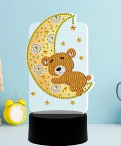 Un encantador Lámpara LED Diamond Painting Kit 5D Oso Durmiente, presenta un soporte negro y una figura de acrílico transparente con un oso marrón decorado con cuentas, abrazado a una luna igualmente adornada y rodeado de estrellas amarillas, acompañado por un reloj despertador amarillo, una figura de modelo de madera y tijeras de mango negro, todo sobre un fondo azul claro.