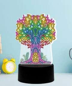 Un árbol decorativo hecho de gemas de colores sobre una base negra, acompañado de un reloj amarillo y un maniquí de madera en un fondo azul claro, junto a tijeras en un soporte blanco, todo centrado en la pieza central: Lámpara LED Diamond Painting Árbol de la Vida.