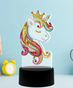 Unicornio de fantasía radiante con crin multicolor, decorado con brillantes gemas y coronado con una diadema dorada, sobre una base negra en un ambiente creativo, titulado 'Lámpara Diamond Painting LED Unicornio 5D'.