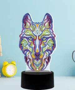 En un fondo azul claro, se exhibe una figura de lobo multicolor decorada con gemas sobre una base negra, junto a un maniquí de madera, un reloj despertador amarillo y tijeras metálicas, destacando la "Lámpara LED Acrílica Lobo Tribal Diamond Paint" frente a una caja blanca con textura cuadrada.