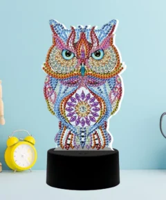En un fondo azul claro, se destaca una "Lámpara LED Búho Místico Bordado Con Diamantes", decorada con coloridas gemas y posada sobre una base negra, acompañada por una figura de maniquí de madera, un reloj despertador amarillo y unas tijeras en un soporte de malla metálica.