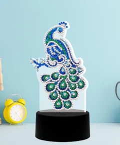 Un maniquí de madera se encuentra al lado de un reloj despertador amarillo y una figura decorativa de pavo real adornada con cuentas azules y verdes sobre una base negra, recuerde que esta "Lámpara LED Pavo Real Pintura de Diamantes" crea un ambiente fascinante junto a unas tijeras en un portatijeras cuadrado contra un fondo azul claro.