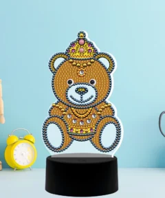Un osito decorado con piedras brillantes y una corona de joyas descansa sobre un soporte negro contra un fondo azul claro, acompañado por un reloj amarillo, una figura de madera articulada y unas tijeras grises en un recipiente cuadrado, creando una escena creativa titulada "Lámpara Rey Oso Punto de Cruz de Diamantes".