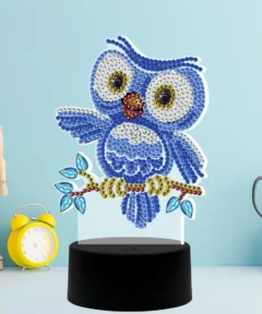 Figura de búho creada con gemas brillantes sobre un soporte negro, acompañada de un reloj despertador amarillo, maniquí de madera, tijeras y una taza amarilla sobre un fondo azul claro, destacando la Lámpara Punto de Cruz de Diamantes Búho 5D.