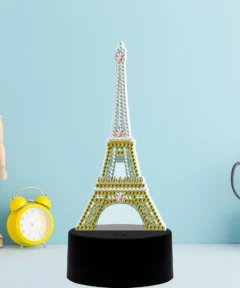 Maniquí de madera junto a un reloj despertador amarillo, acompañados de una Lámpara LED Torre Eiffel Punto de Diamante sobre base negra, con tijeras y un recipiente de malla lleno de lapiceros, todo sobre un fondo azul claro.