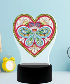 Un diseño vibrante de mariposa en el centro de una figura de corazón, adornado con piedras brillantes de colores, se destaca sobre una base negra, acompañado por un reloj despertador amarillo, una figura de maniquí de madera y tijeras en un portatijeras, todo sobre un fondo azul claro, evocando la Lámpara Corazón Mágico 5D Pintura Diamante.