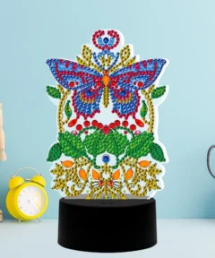 Modelo de mariposa decorado con gemas de colores brillantes sobre un fondo azul claro, presentado como una Lámpara Decorativa Diamond Painting Mariposa, acompañado de un reloj despertador amarillo y una figura de madera articulada, todo sobre una base negra circular junto a unas tijeras plateadas en un recipiente y un cuaderno con espiral.