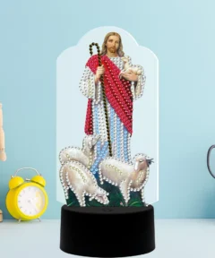 Figura mágica sosteniendo un cordero y un bastón, envuelta en un manto rojo con decoraciones brillantes, flanqueada por tres ovejas decoradas en la base negra, todo sobre un fondo azul junto a un reloj despertador amarillo, un maniquí de madera y tijeras en un cubilete, destacando el tema de 'Corazón Brillante Lámpara LED Diamond Painting'.