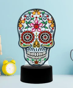 Calavera decorativa con pedrería de colores sobre una base negra, acompañada de una figura de madera articulada, reloj despertador amarillo y tijeras plateadas en un recipiente blanco, todo dispuesto sobre un fondo azul, destacando como un Corazón Brillante Lámpara LED Diamond Painting.