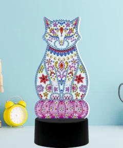 Una figura decorativa de un gato adornado con gemas de colores destaca sobre una base negra circular al lado de un reloj despertador amarillo, un maniquí de madera y unas tijeras en un soporte, todo enmarcado por un fondo azul claro, y complementado por el elegante diseño de la Lámpara LED Gato Mandala Punto De Diamante 5D.