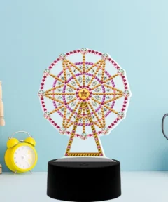 Modelo de una noria decorada con piedras de colores sobre una base negra, acompañado de un reloj despertador amarillo, un maniquí de madera articulado y unas tijeras en un soporte blanco, todo en un fondo azul claro, titulado 'Lámpara Decorativa Noria Pintura de Diamante'.