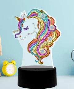 Unicornio decorado con gemas de colores brillantes sobre una base negra, contrastando con un fondo azul claro, acompañado de una figura de madera articulada, un reloj amarillo, tijeras, y una caja blanca con diseño de cuadrícula, presentado como una 'Lámpara de Unicornio Arcoíris Diamond Painting'.