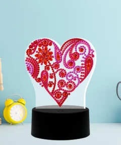 Corazón Brillante Lámpara LED Diamond Painting con gemas rojas y rosadas sobre una base circular negra, acompañado de un maniquí de madera, un reloj despertador amarillo y unas tijeras en soporte, todo en un fondo azul claro con un bloc de notas.