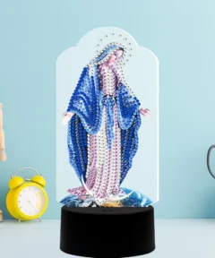 Una figura mágica con aspecto religioso, adornada con gemas brillantes en un fondo azul claro, se alza sobre una base negra junto a un reloj despertador amarillo y un maniquí de madera, complementada con tijeras en un soporte, destacando la presencia de un "Corazón Brillante Lámpara LED Diamond Painting".