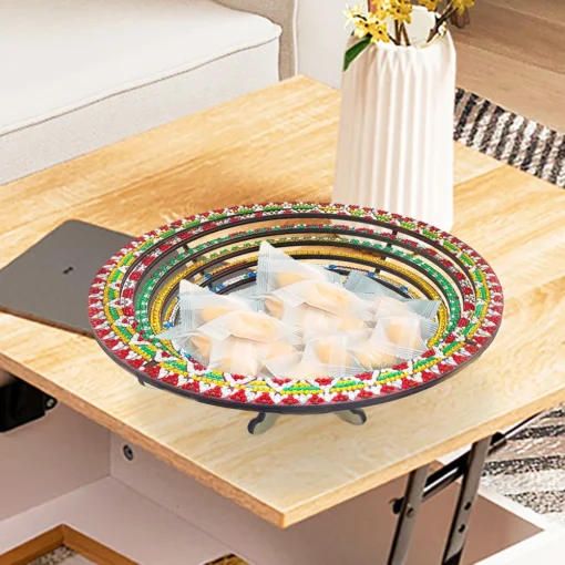 En una mesa de madera, destaca un Plato Frutero Mandala Diamond Painting DIY con bordes coloridos lleno de dulces envueltos en plástico transparente, acompañado por un sofá al fondo, un jarrón blanco con flores amarillas y una alfombra rayada en blanco y negro.