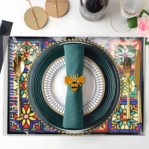 En la elegante mesa se aprecia un Mantel Pintura de Diamantes Artísticos 5D Antideslizantes con diseño floral colorido, que acompaña un plato grande oscuro con borde dorado, un plato pequeño con patrón en blanco y negro, una servilleta verde adornada con un anillo dorado en forma de abeja, cubiertos dorados, una copa de vidrio con borde dorado, una botella de vidrio, una vela dorada, y una flor rosa en la parte superior derecha.