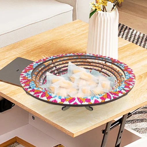 Sobre una mesa de madera, un Plato Decorativo Cocina Punto de Diamantes adornado con cuentas de colores contiene múltiples paquetes transparentes, mientras que al fondo se observa un jarrón blanco con flores amarillas y verdes junto a un sofá de color claro.