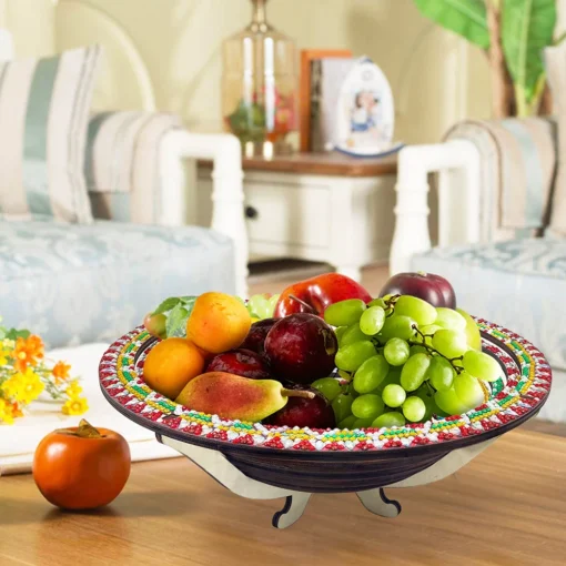 Frutero con diseño colorido que contiene un racimo de uvas verdes, duraznos, higos y manzanas, situado sobre una mesa en una sala de estar elegante con sofás blancos, cojines a rayas, mesa de centro decorada con libro, jarrón y flores amarillas, con un kaki adicional sobre la mesa, representando un 'Plato Decorativo Cocina Punto de Diamantes'.